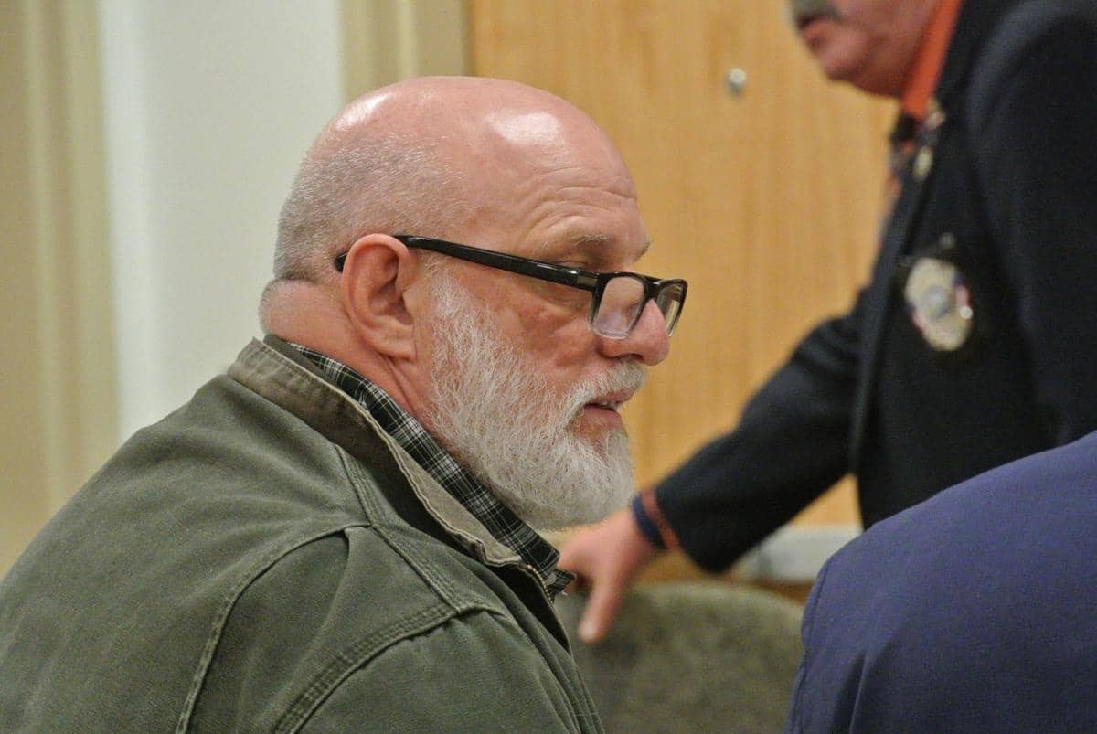 A bald man with glasses and a beard sitting in a courtroom.